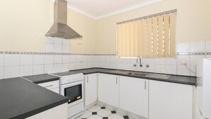 Una cocina con gabinetes blancos, encimera negra, estufa, pileta y ventana con persianas.