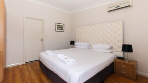 Una cama bien hecha con sábanas blancas y dos almohadas, una mesita de noche con lámpara y un aire acondicionado de pared.