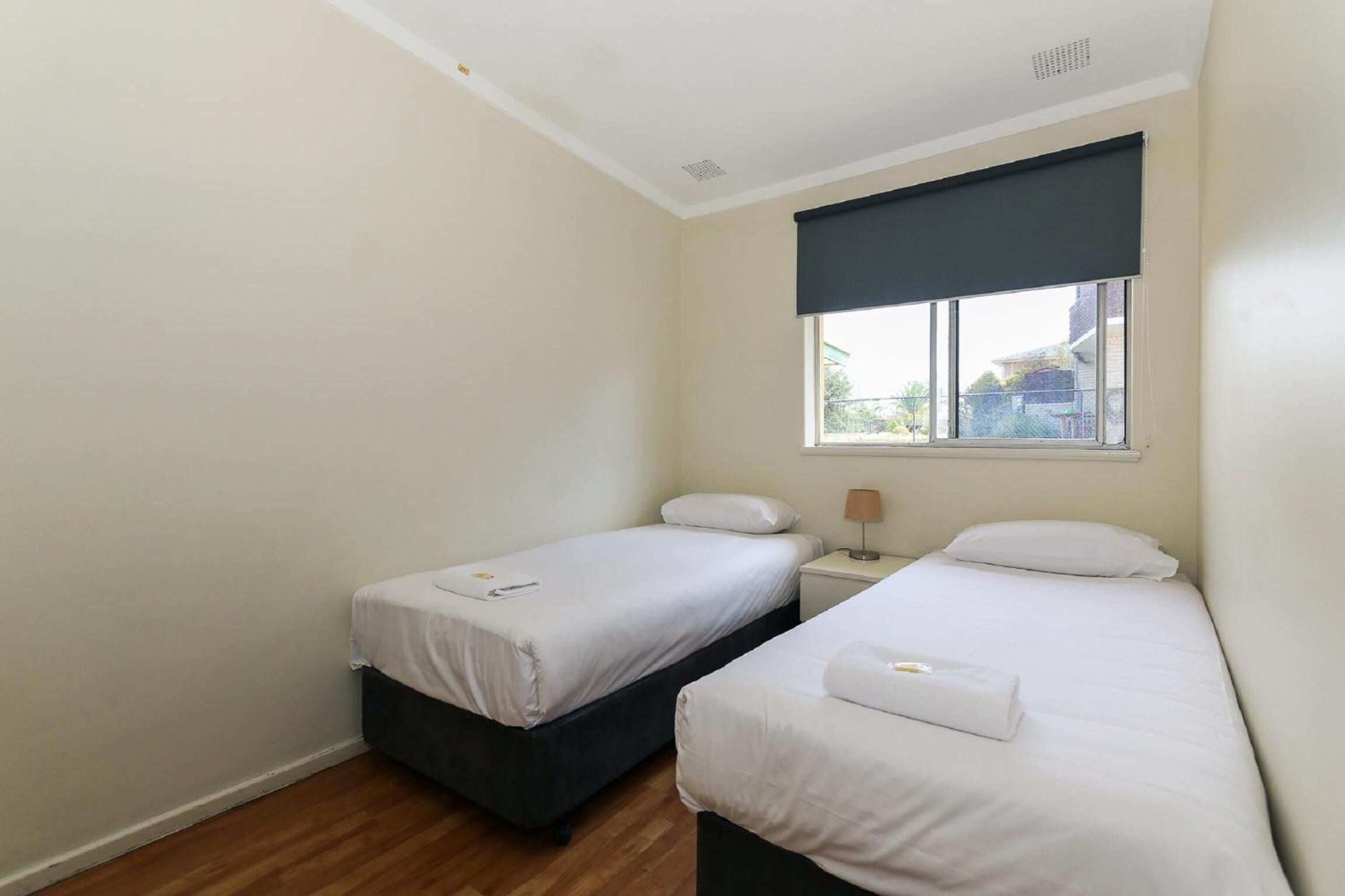 A small, clean room with two single beds, a window with a dark blind, and a small bedside table with a lamp.