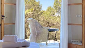 Una stanza luminosa con una poltrona in vimini, un tavolino, un asciugamano bianco arrotolato e vista su alberi e cielo.