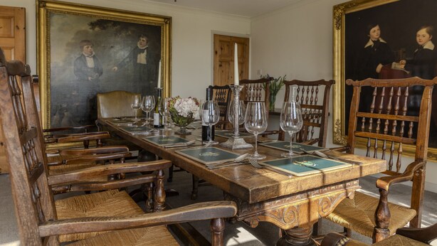 Une salle à manger élégante, avec une longue table en bois dressée pour six personnes, entourée de chaises en bois. Les murs sont ornés de tableaux, un lustre illumine la pièce et un vase rempli de fleurs est posé sur la table.