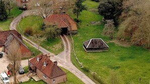 Vue aérienne d’une zone rurale avec une grande maison, un bâtiment plus petit et une grange. Des routes relient les bâtiments, et le paysage est entouré de champs et d’arbres.