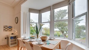 A dining area with a wooden table, white chairs, and a view of trees through large windows. - 3 Bed in Mortehoe (oc-pines) (Mortehoe)