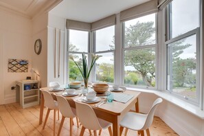 Ein Essbereich mit Holztisch, weißen Stühlen und Blick auf Bäume durch große Fenster.