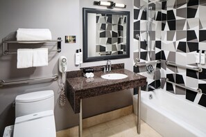 Un baño moderno con lavamanos de encimera de granito, inodoro blanco, bañera con ducha y pared con estampado geométrico.