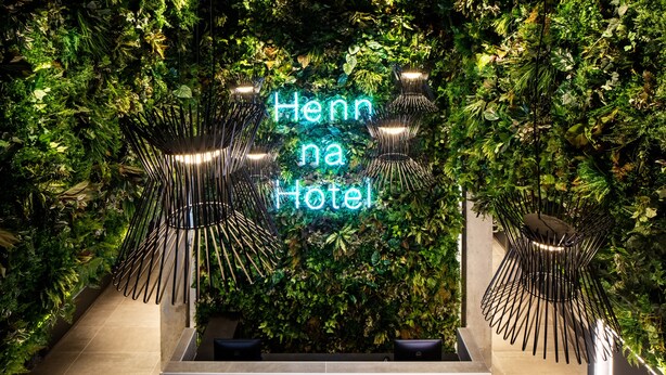Un espace d’accueil agrémenté d’un panneau néon indiquant « Henn na Hotel », entouré d’une végétation luxuriante et de luminaires modernes.