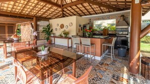 A covered outdoor dining area with a glass table and wicker chairs, a kitchen with a grill, and decorative plates on the wall. - GB22 Ótimo Apt 3 Quartos - Guarajuba (Camaçari)