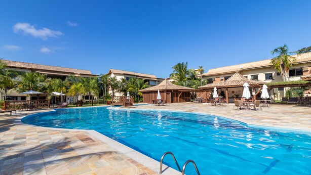 A swimming pool with a tiled deck, lounge chairs, umbrellas, and palm trees. - GB22 Ótimo Apt 3 Quartos - Guarajuba (Camaçari)