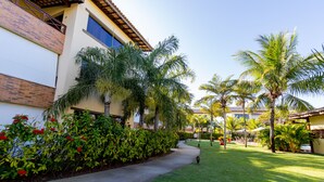 A courtyard with palm trees, a pathway, and a building with a tiled roof and brick facade. - GB22 Ótimo Apt 3 Quartos - Guarajuba (Camaçari)