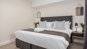 Ein ordentlich bezogenes Bett mit grauem Kopfteil, weißer Bettwäsche und gemusterten Kissen. Ein kleiner Nachttisch mit Lampe und Topfpflanze steht daneben. Der Raum ist minimalistisch eingerichtet, mit hellen Wänden und einer Deckenleuchte.