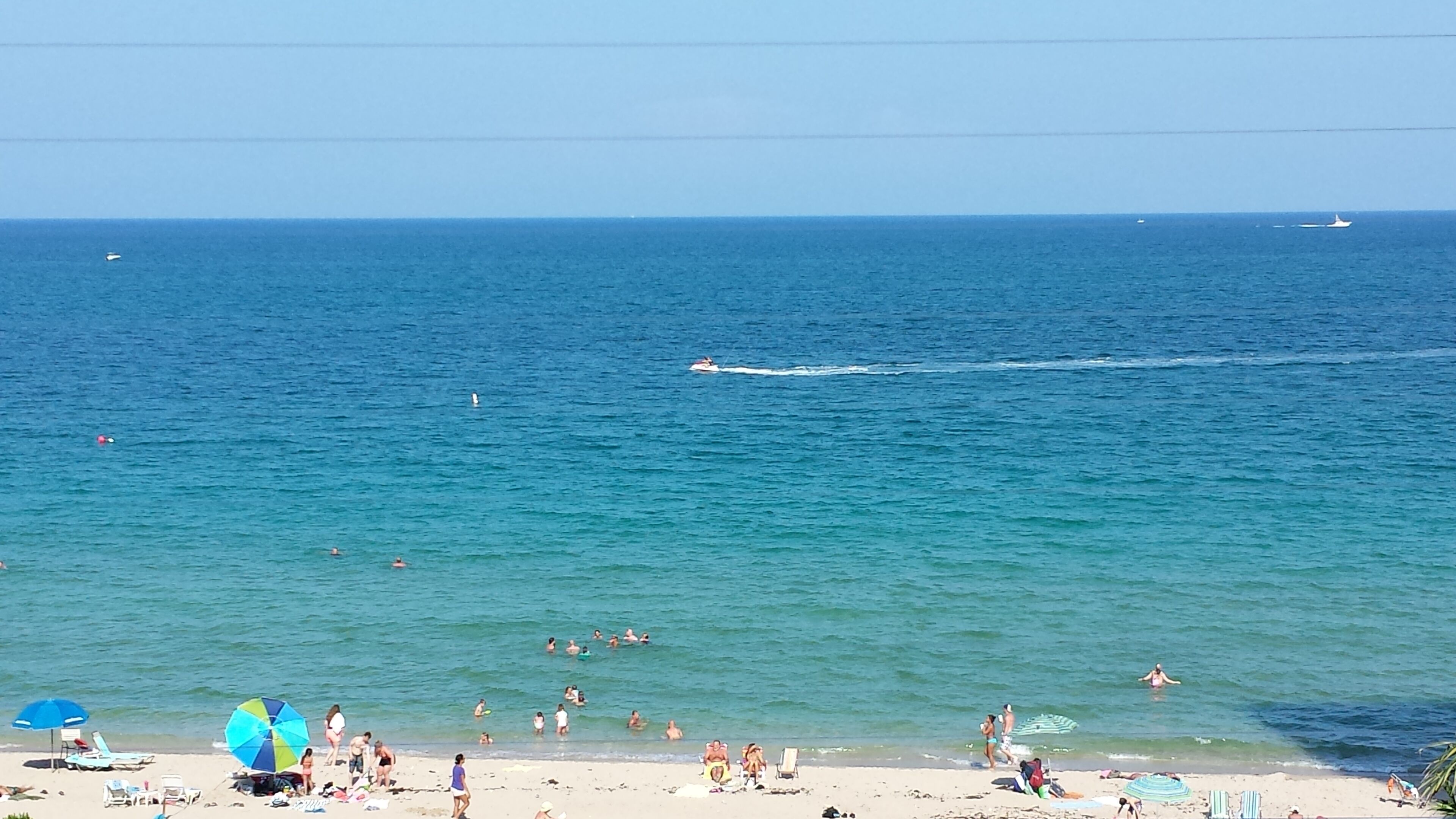 Una playa con gente nadando y tomando el sol, un bote en el agua, y un cielo despejado.