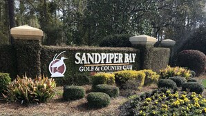 Sandpiper Bay Golf & Country Club -kyltti, jossa on logon kuva, ja sen ympärillä on hoidettuja puutarhoja ja siististi leikattuja pensasaitoja.