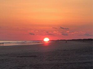 Een strand bij zonsondergang, met de zon laag aan de horizon, mensen wandelen langs de kust en een heldere lucht.