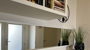 Un interior modern amb un mirall gran, una prestatgeria plena de llibres i dues plantes en testos sobre una superfície.