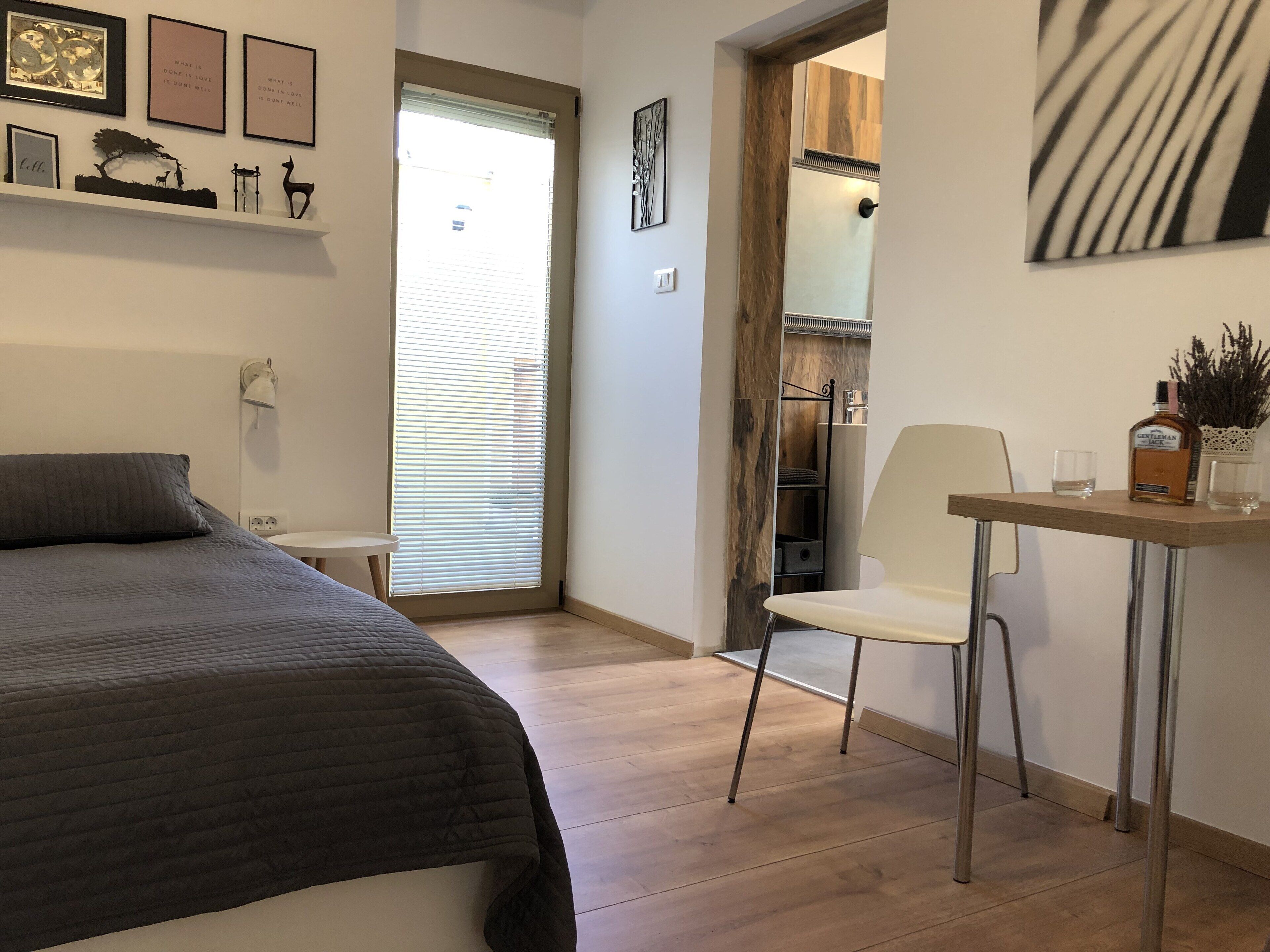 Une chambre à coucher avec un lit, une chaise, une table sur laquelle se trouvent une bouteille et des verres, et une fenêtre avec des stores.