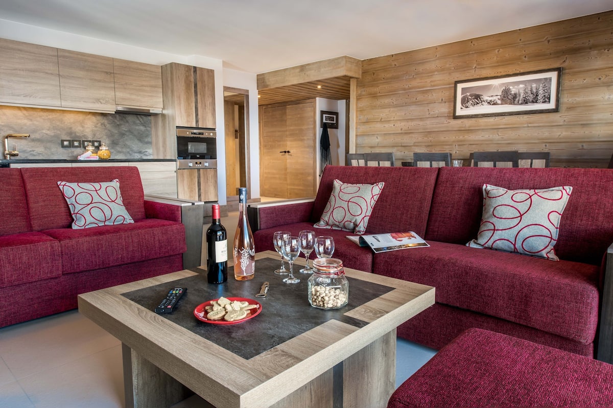 A modern living room with a red sofa set, a coffee table with remote control, wine bottle, glasses, and snacks.