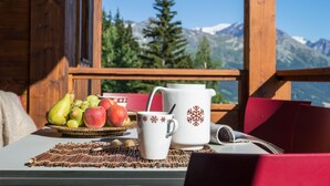 Una mesa con una cesta de frutas, una taza blanca y una jarra blanca con diseño de copo de nieve, frente a un paisaje montañoso.