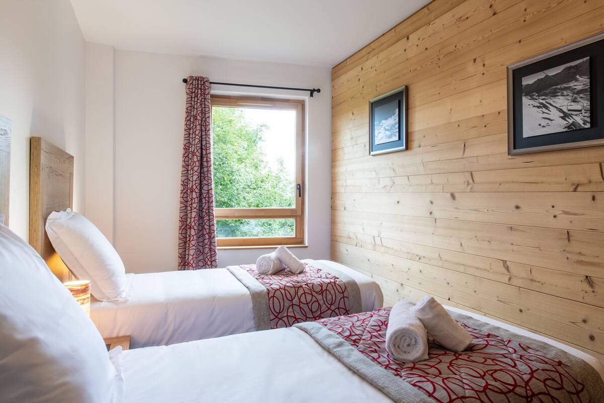 A hotel room with two beds, a window with a view of greenery, wooden paneling on the walls, and a framed picture.