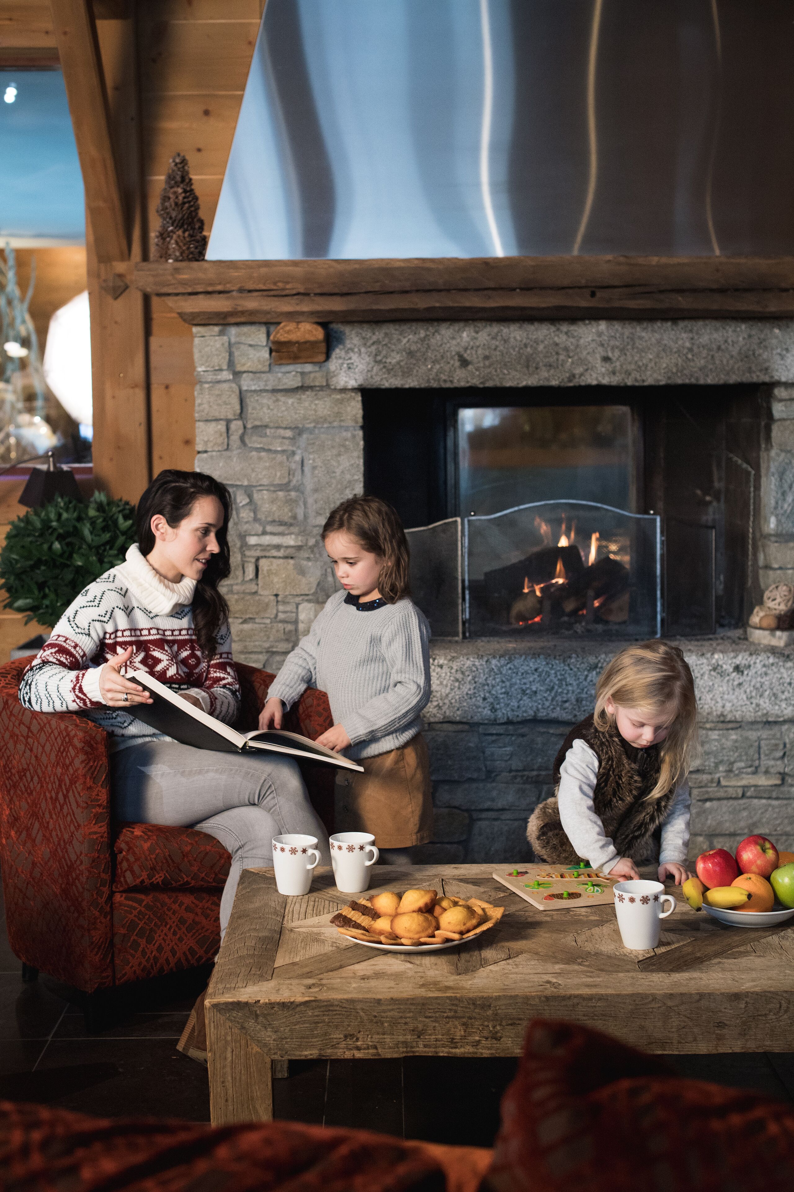 Un salon confortable avec une cheminée, une table basse sur laquelle sont disposés des en-cas, et une personne qui lit un livre.