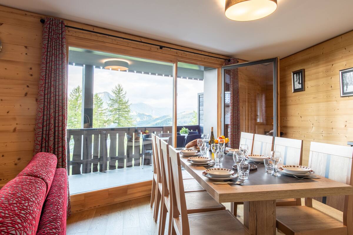 A dining area with a wooden table set for four, a bench, and a view of mountains through large windows.