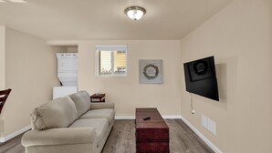 Een woonkamer met een beige bank, een flatscreen televisie aan de muur, een kleine houten tafel en een raam met zonwering.