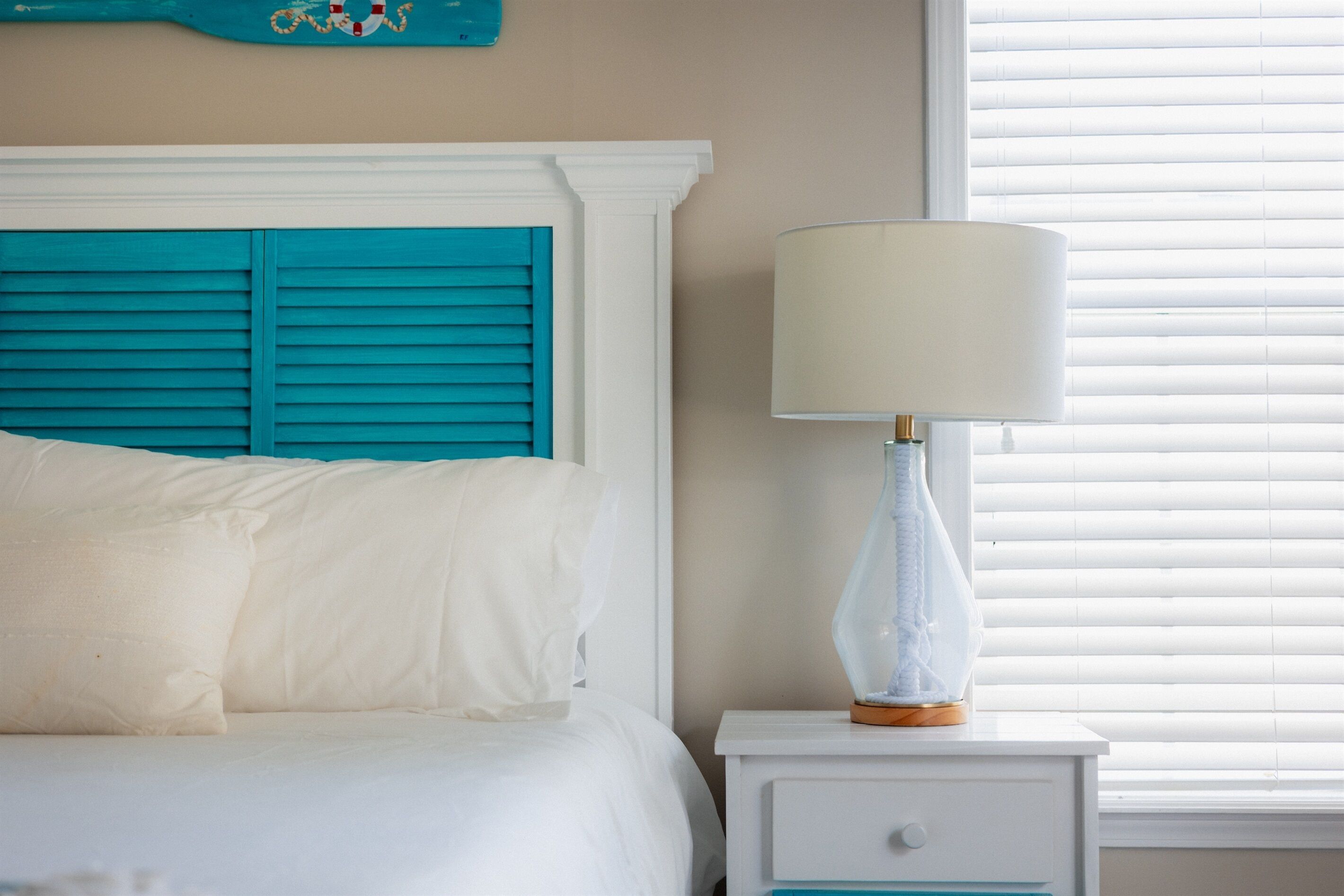 A bed with white bedding and a teal headboard, a white nightstand with a lamp, and a window with blinds.