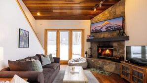 A cozy living room with a stone fireplace, a large flat-screen TV mounted above it, and a comfortable sofa. - 100 Steps Away, Ski in Ski Out Condo (Big Sky)