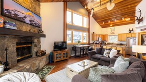 A cozy living room with a fireplace, a television, and a dining area with a kitchen in the background. - 100 Steps Away, Ski in Ski Out Condo (Big Sky)