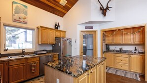 A kitchen with wooden cabinets, granite countertops, a stainless steel refrigerator, and a deer head mounted on the wall. - 100 Steps Away, Ski in Ski Out Condo (Big Sky)