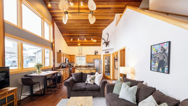 A spacious living room with a large brown sofa, a wooden coffee table, and a TV on a wooden cabinet. There is a kitchen in the background with wooden cabinets and a large window. - 100 Steps Away, Ski in Ski Out Condo (Big Sky)