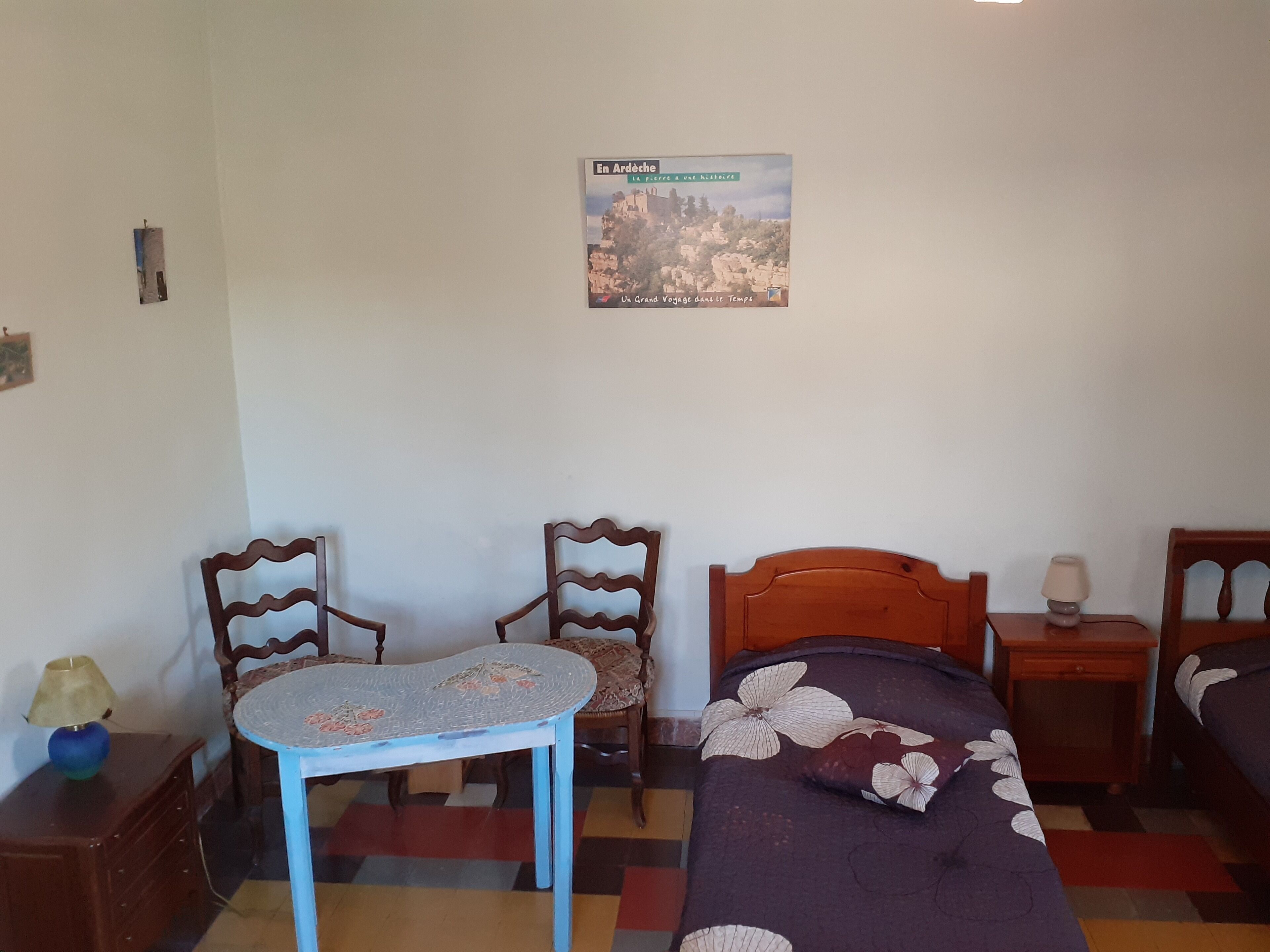 Una habitació amb un llit, una taula petita, dues cadires, una tauleta de nit i un cartell a la paret.