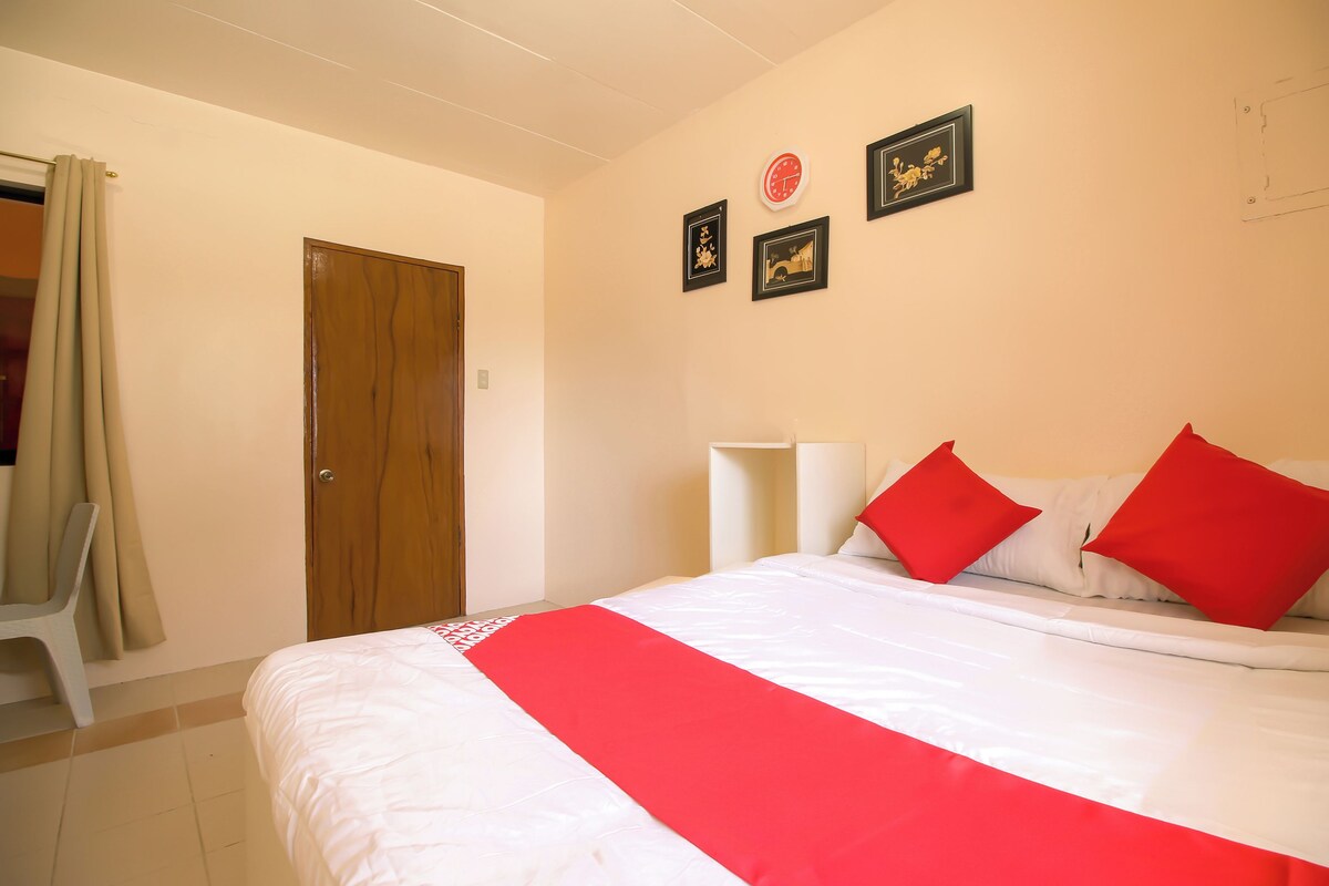 A hotel room with a bed, a wooden door, a white chair, and a wall with framed pictures.