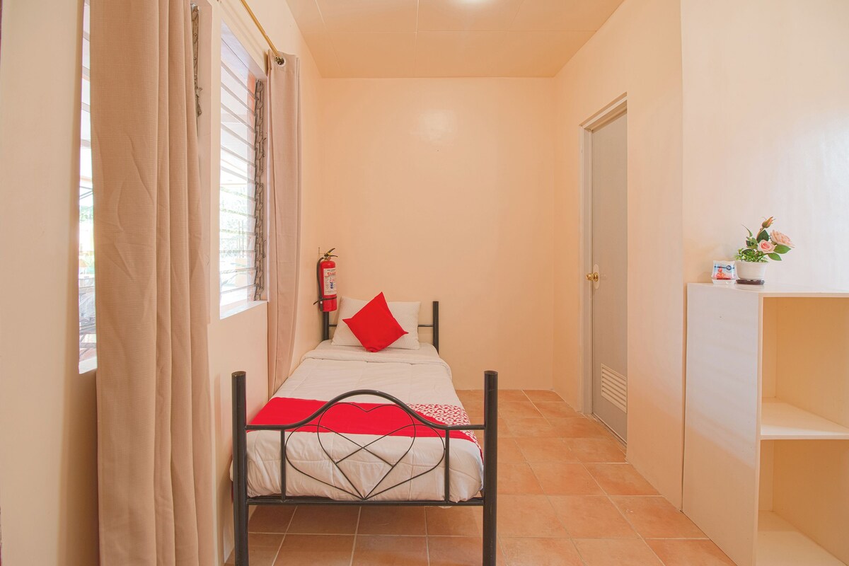 Una stanza piccola con un letto, un estintore, una porta e un piccolo ripiano con una pianta.