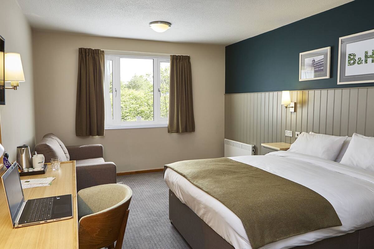 A hotel room with a bed, a desk with a laptop, a chair, a window with curtains, and a wall-mounted lamp.