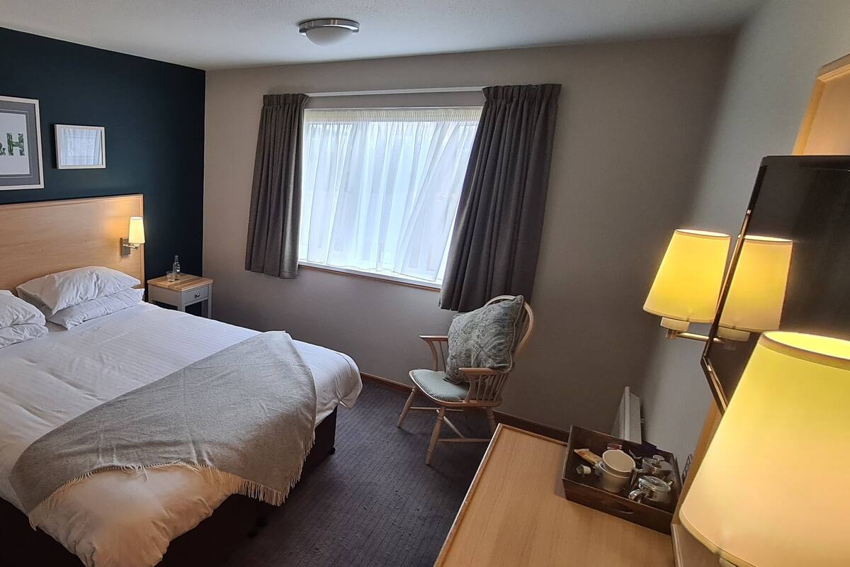 A hotel room with a large bed, two bedside tables, a chair, a window with curtains, and framed pictures on the wall.