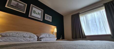 A hotel room with a bed, two framed pictures on the wall, a nightstand with a lamp, and a window with curtains.