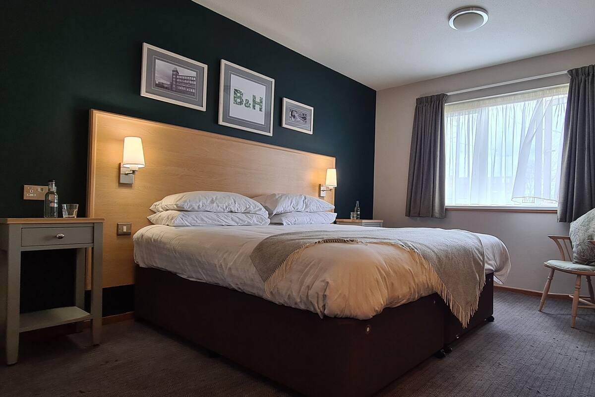 A hotel room with a bed, bedside tables, a chair, a desk, a window with curtains, and wall-mounted artwork.