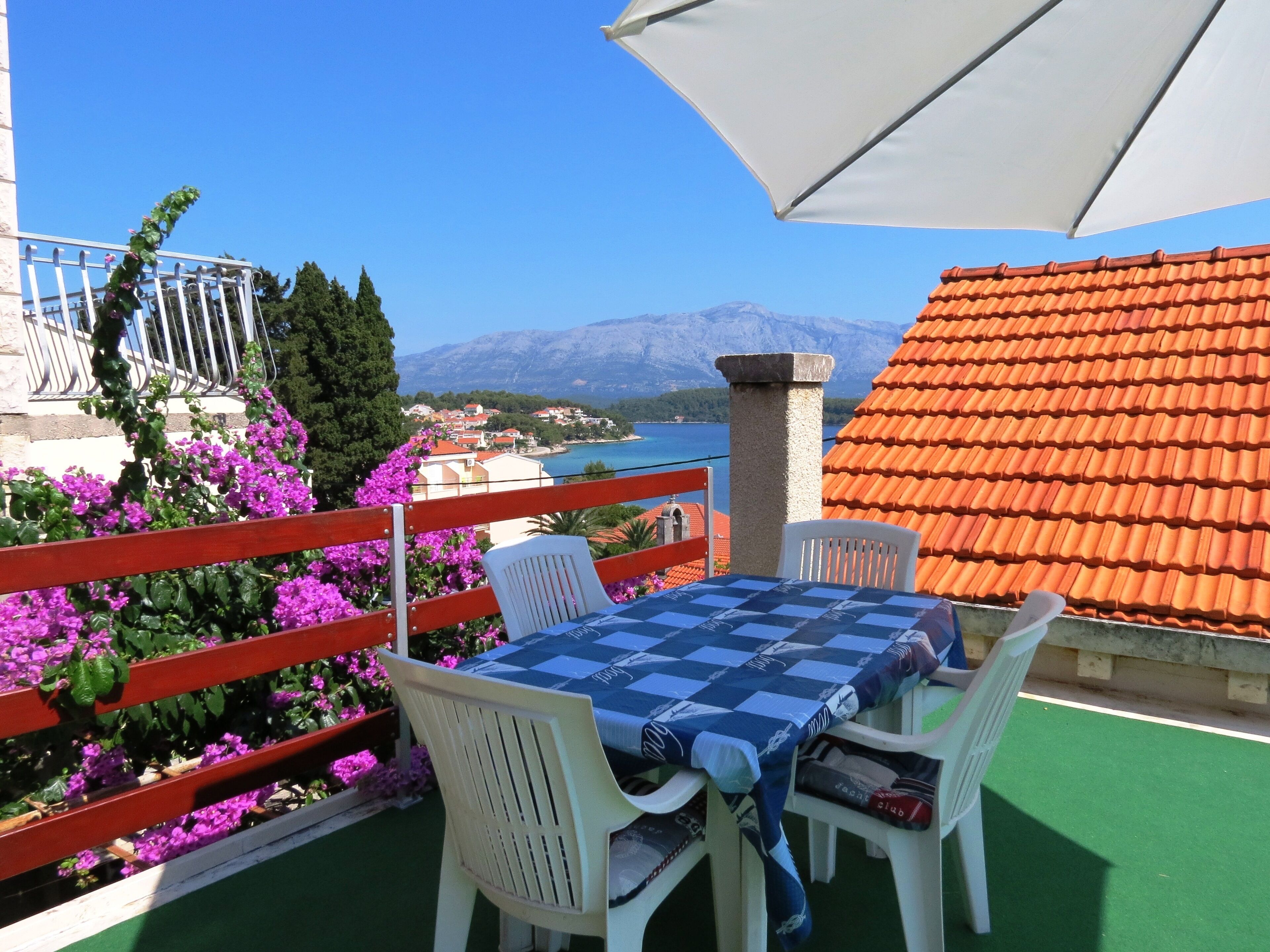 Um terraço com toalha de mesa xadrez, cadeiras brancas, corrimão vermelho e flores roxas.