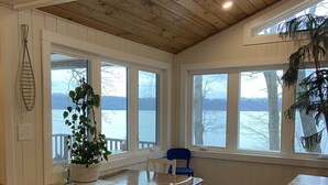 Un espace repas avec une table et des chaises en bois, de grandes fenêtres donnant sur l’eau et un plafond avec des poutres apparentes.