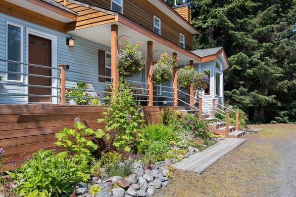 Une maison en bois à deux étages, dotée d’un porche couvert, d’un chemin en pierre et agrémentée de diverses plantes et fleurs.