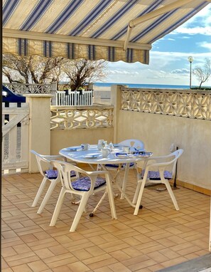 Een buitenterras met een ronde tafel en witte plastic stoelen, een overdekt gebied met een gestreept dak en uitzicht op zee.