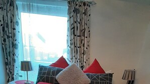 Un lit bien fait, avec une literie à motifs géométriques et plusieurs oreillers, dont certains rouges et blancs. Il y a deux tables de chevet avec des lampes, et la pièce est dotée d’une fenêtre avec des rideaux.