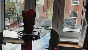Une pièce avec une table en verre, un vase rouge et une vue sur un bâtiment en briques, visible à travers une porte-fenêtre coulissante.