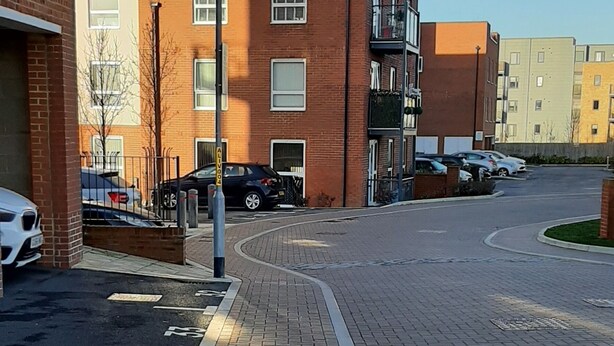 Un trottoir dallé bordé d’une bordure, situé à côté d’un immeuble à plusieurs étages dont la façade est en briques, avec un lampadaire et des voitures garées.