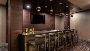 Uma área de bar moderna com um balcão extenso, vários banquinhos, iluminação pendente e uma televisão de tela plana na parede.