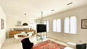 Un salon moderne doté d’un grand téléviseur, d’un canapé, d’un tapis à motifs rouges et d’un coin repas avec une table et des chaises blanches.