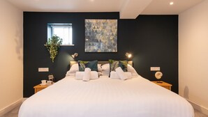 Ein modernes Schlafzimmer mit einem großen Bett, einer geometrischen Wanddekoration, einem Fenster mit Vorhängen und einem kleinen Tisch mit Tasse und dekorativem Gegenstand.
