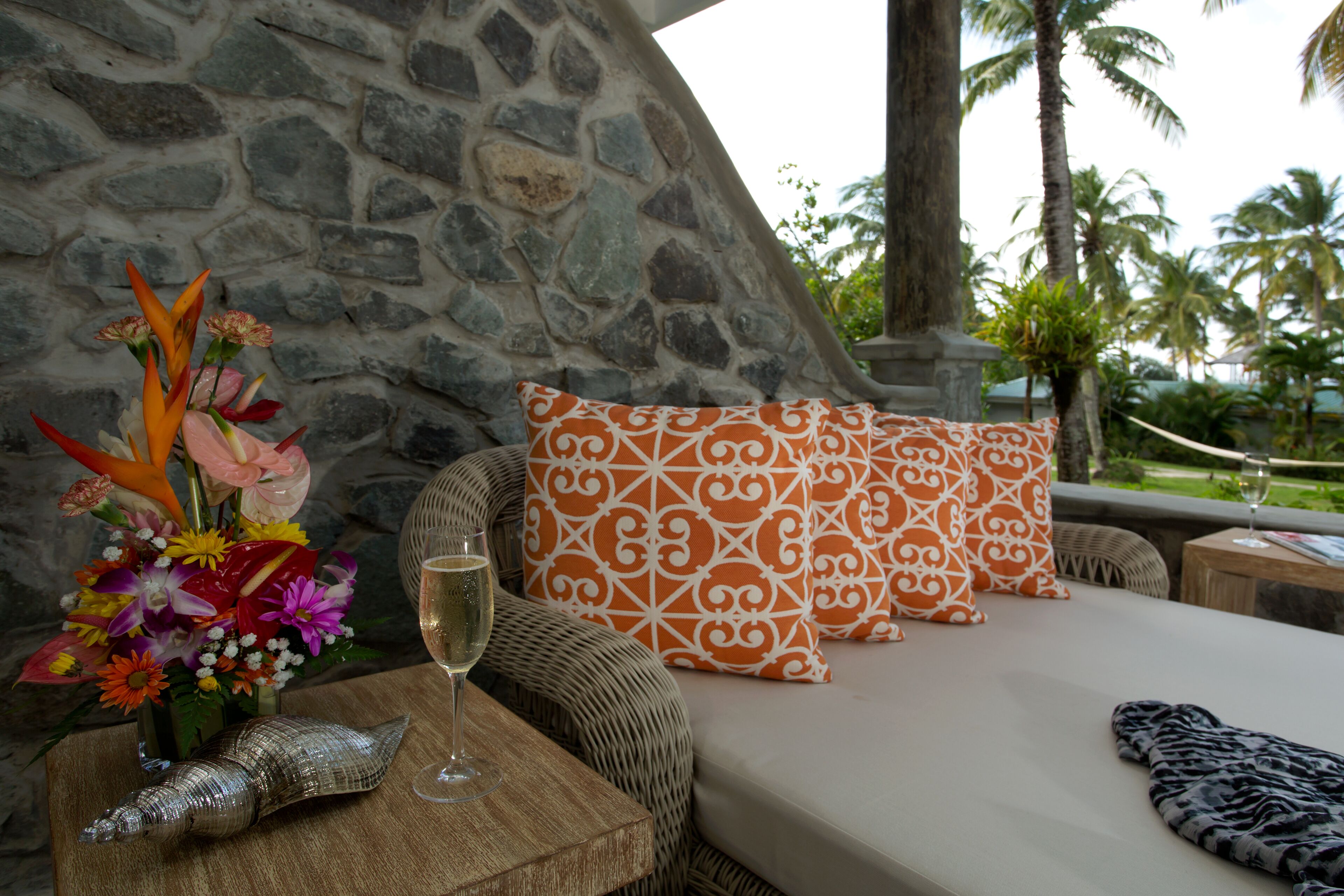 Una habitación con pared de piedra, cama con almohadas estampadas, sofá de mimbre, una mesita con una copa de vino y una concha decorativa.