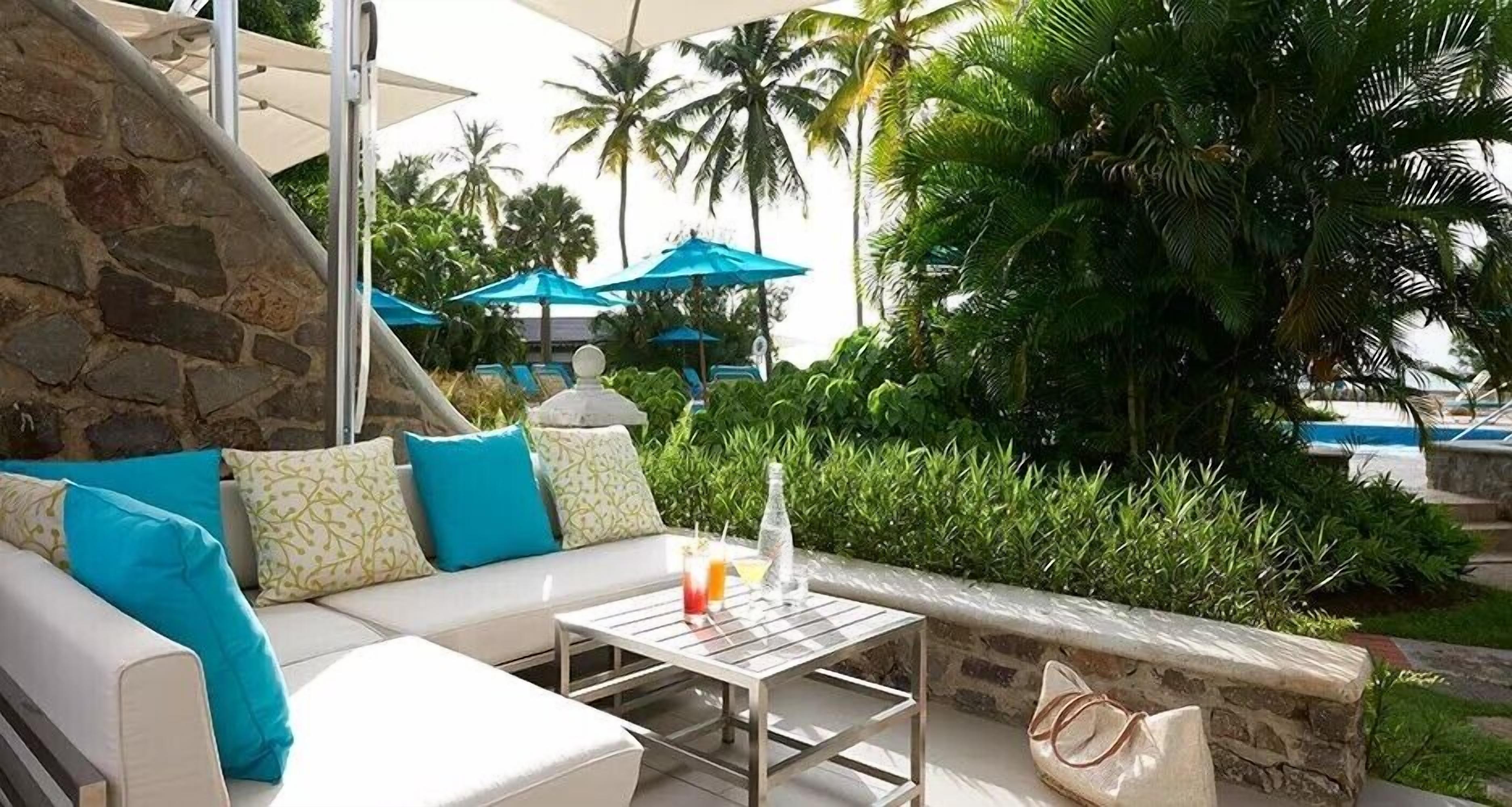 A sunlit outdoor seating area with a stone wall, a white sofa with blue cushions, and a table with a drink.