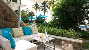 A sunlit outdoor seating area with a stone wall, a white sofa with blue cushions, and a table with a drink.